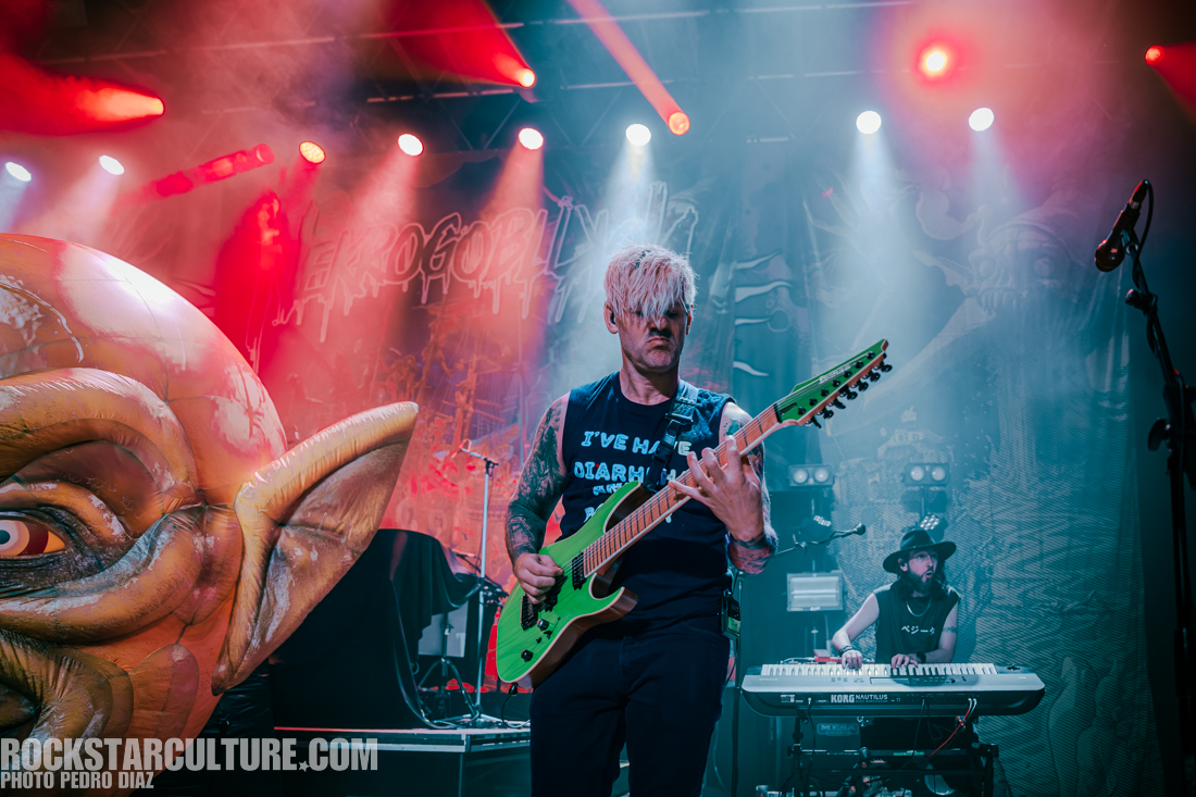 A musician playing an electric guitar on stage with colorful lighting, featuring a large inflatable character in the foreground and another band member playing a keyboard in the background.