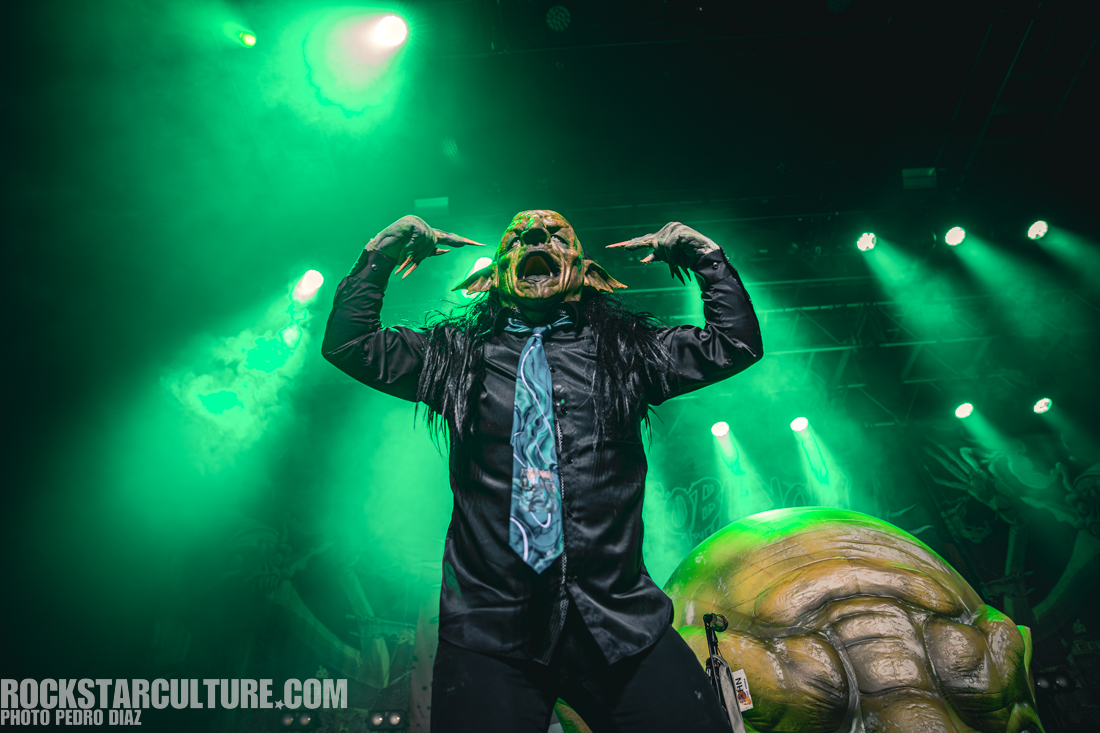 A performer with a grotesque mask and long black hair strikes a dramatic pose on stage, wearing a black shirt and a colorful tie, surrounded by green stage lights and smoke.