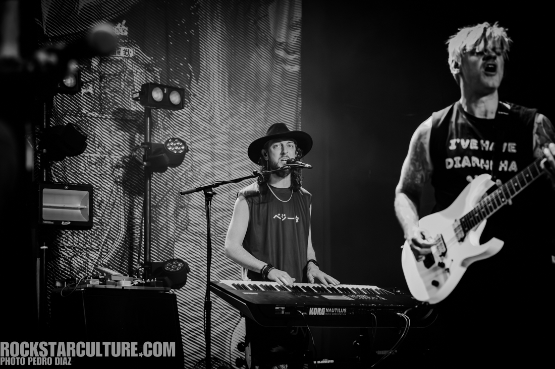 A black and white image of a musician playing a keyboard on stage, wearing a hat and a tank top, while another musician with a guitar is singing beside him. Stage lights are visible in the background.