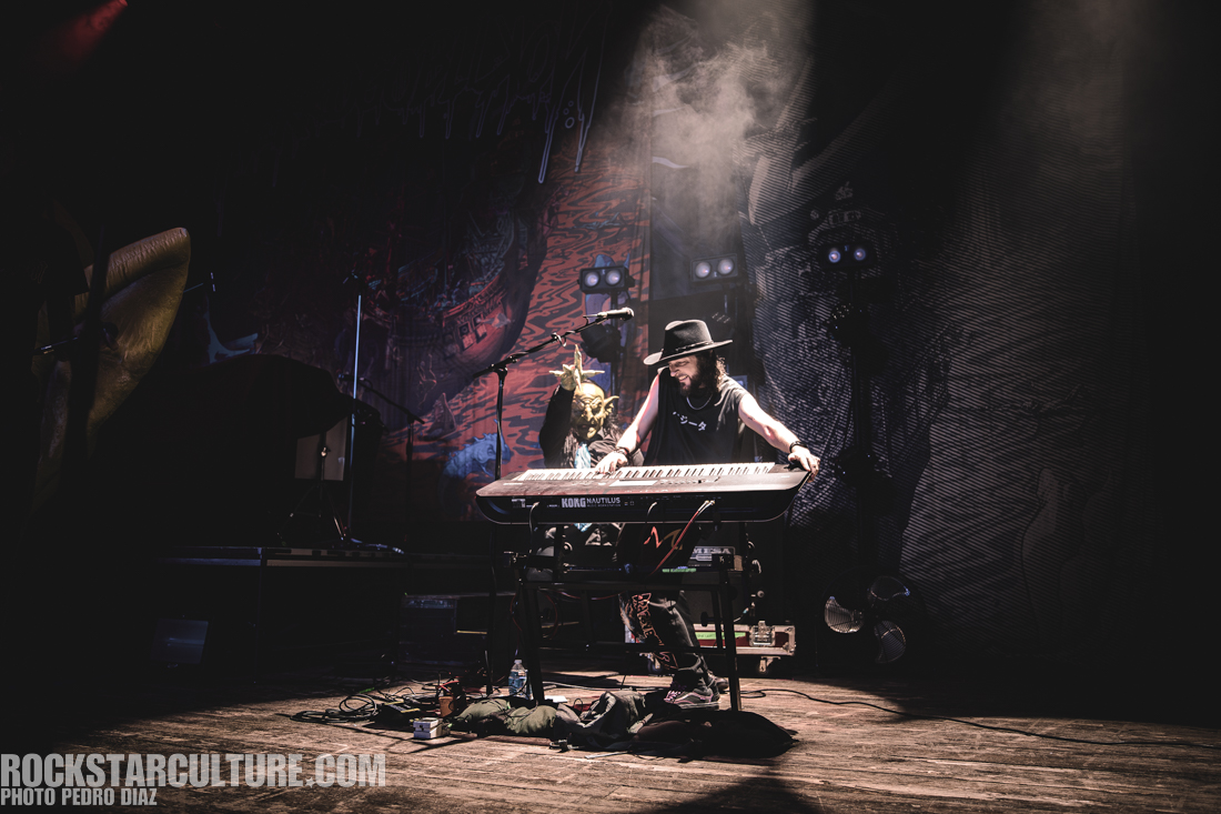 A musician playing a keyboard on stage, wearing a black hat and casual clothing, with a dramatic backdrop and stage lighting.