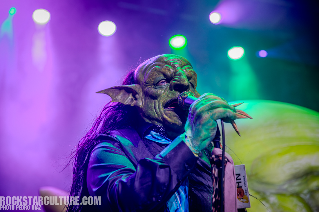 A performer wearing an elaborate green goblin costume singing into a microphone on stage with colorful lighting in the background.