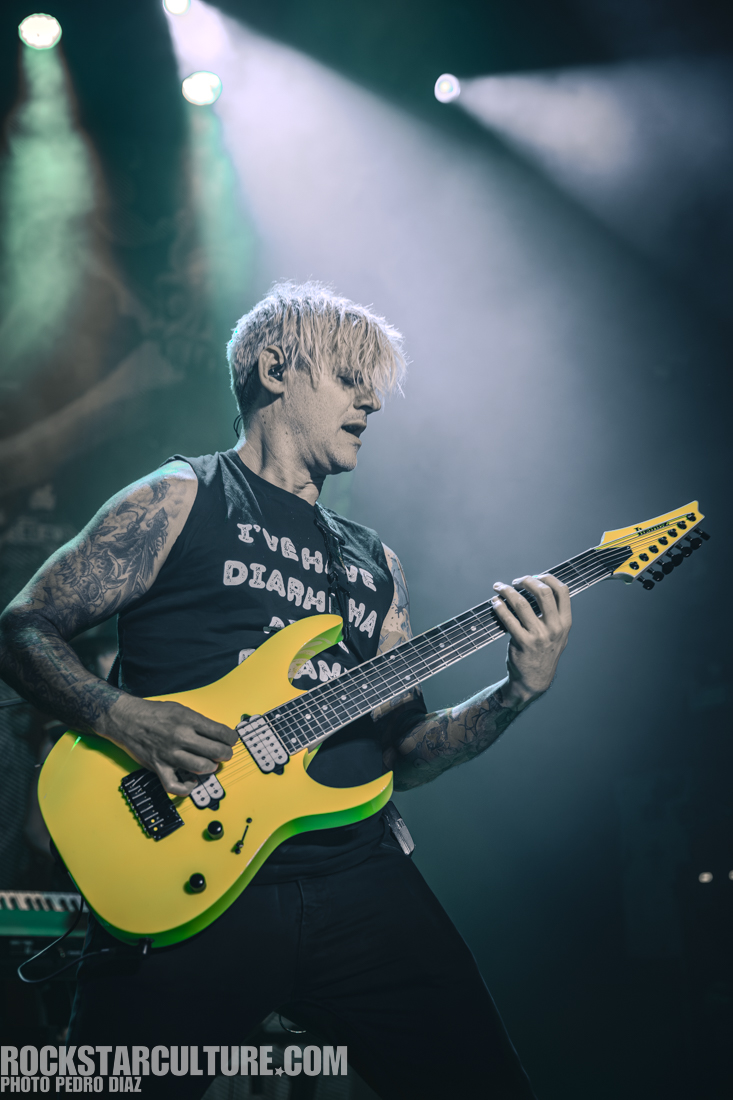 A musician with bleached hair performs on stage, playing a bright yellow guitar, wearing a black tank top with text. The background features stage lights and an energetic atmosphere.