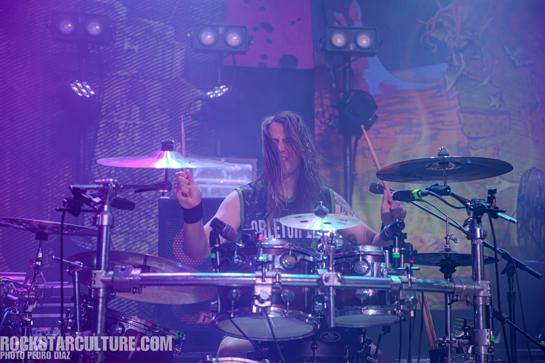 A drummer performing on stage with intense focus, surrounded by colorful stage lights and a vibrant backdrop.