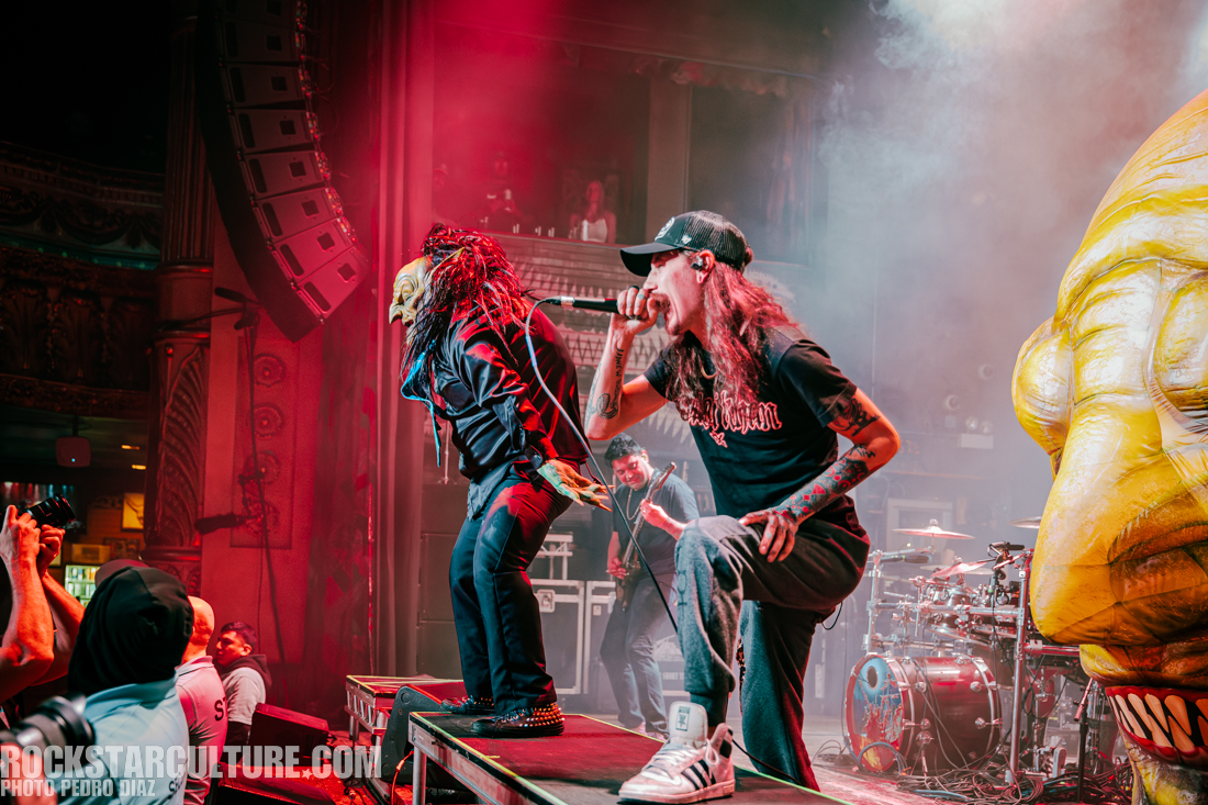 Two male performers on stage during a live music concert, one wearing a mask and the other with long hair, engaging with the audience. Dramatic lighting and a large carved face prop visible in the scene.