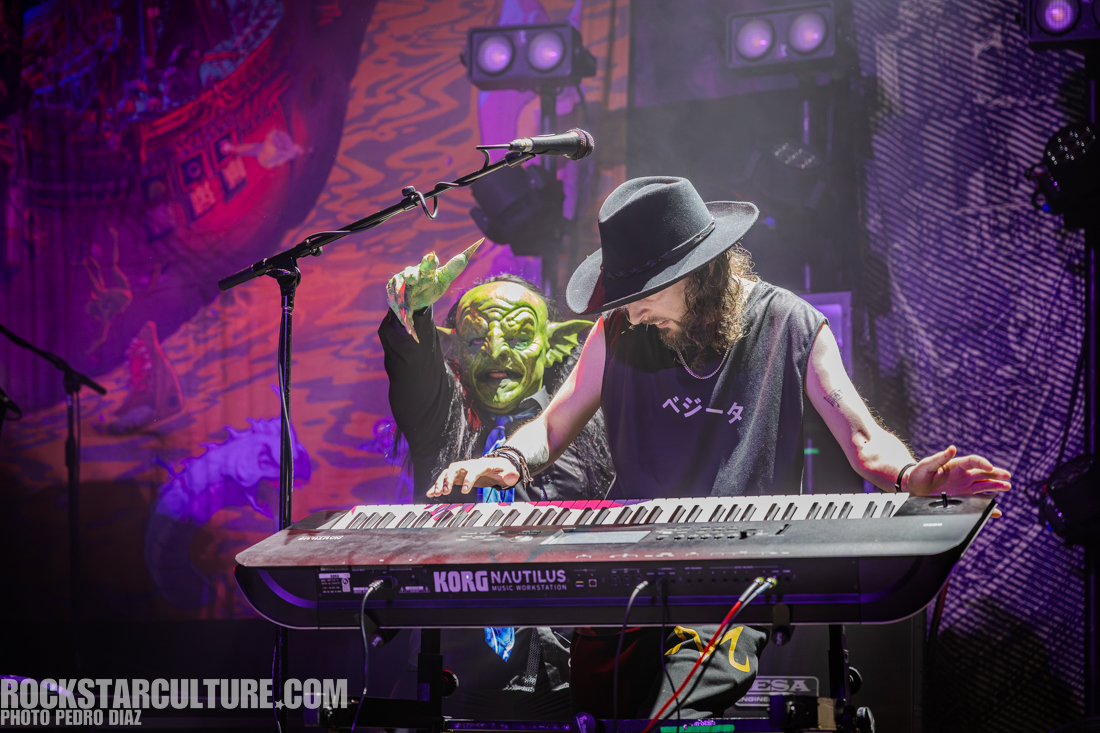 A musician playing a keyboard on stage, with a green, goblin-like character appearing behind them, pointing and gesturing. The stage is lit with colorful lights and a vibrant backdrop.