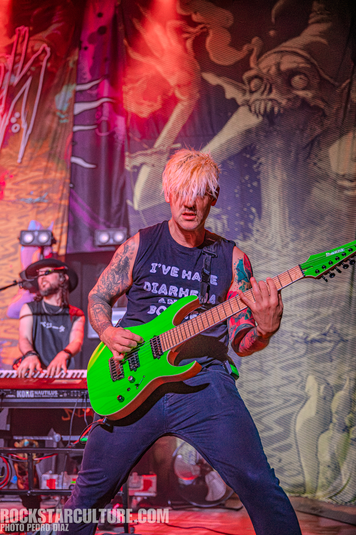 A musician passionately playing a bright green electric guitar during a live performance, with colorful stage lighting and a dynamic background art featuring surreal imagery.