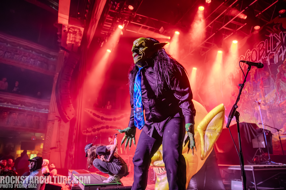A performer dressed in a goblin costume with a green face and long black hair, wearing a black shirt and blue tie, engages with the audience on stage. The background features colorful stage lighting and a large goblin figure.