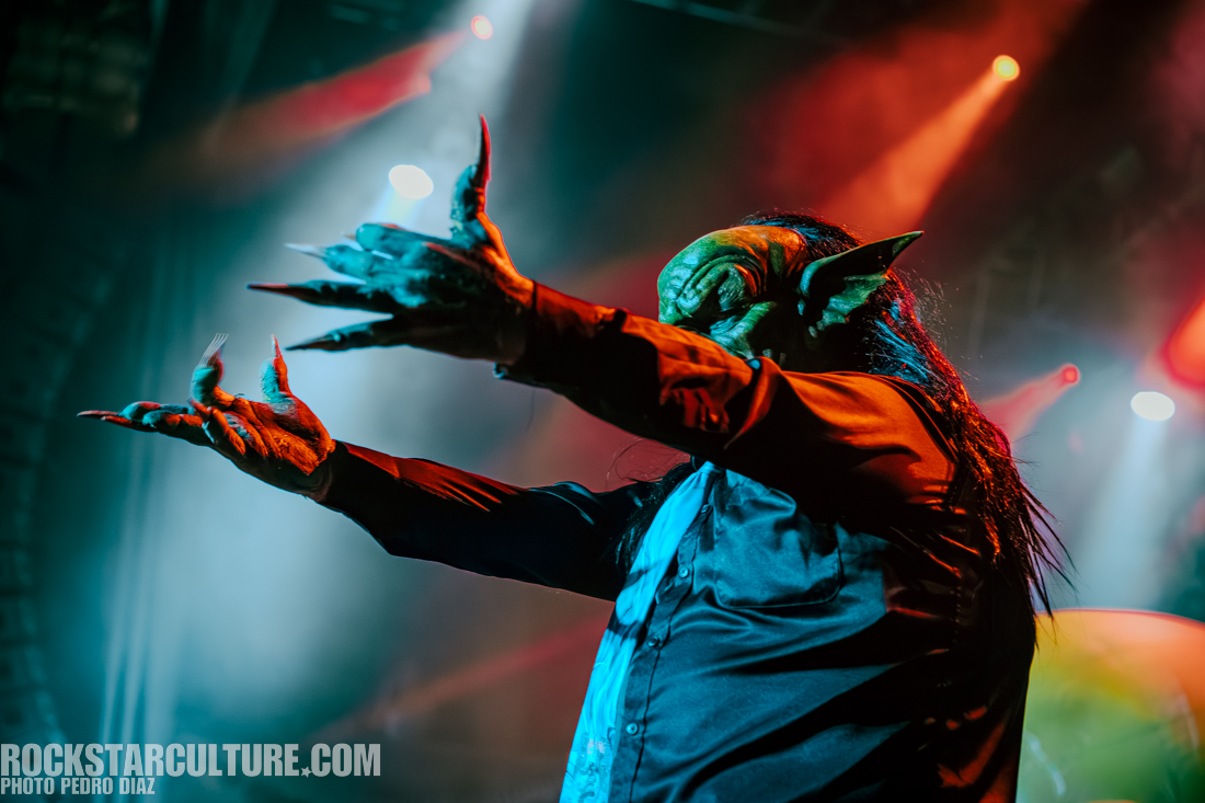 A performer dressed in an elaborate green creature costume with long claws, striking a dramatic pose on stage under colorful lighting.