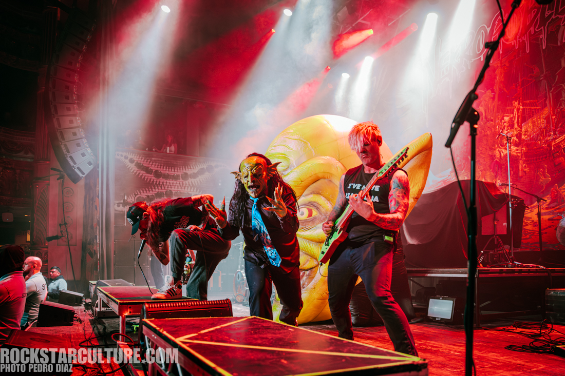 A live music performance featuring three musicians on stage, with one wearing a mask and striking a dramatic pose. The background has vibrant lighting and a large cartoonish face. The atmosphere is energetic and lively.