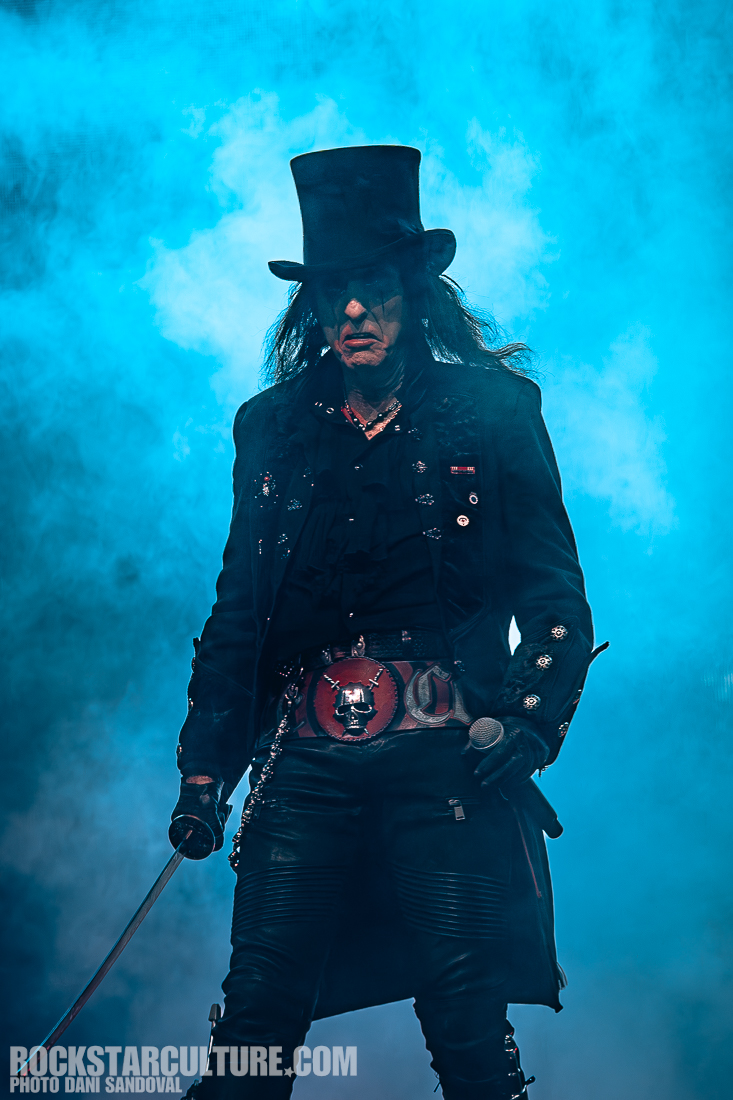 A musician in a black coat and top hat stands confidently on stage, holding a microphone in one hand, surrounded by blue smoke.