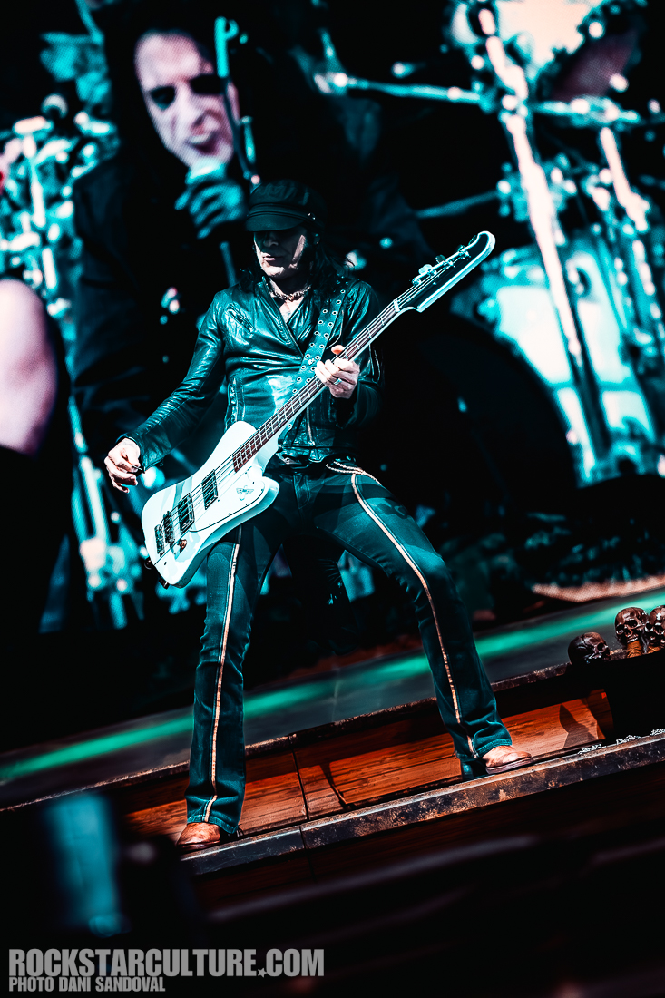 A musician performing on stage, dressed in a leather jacket and pants with vertical stripes, playing a white bass guitar. In the background, blurred images of band members are visible, creating a dynamic concert atmosphere.