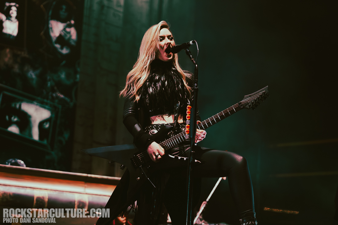 A female guitarist is performing passionately on stage, singing into a microphone while holding a black electric guitar. She has long blonde hair and is wearing a black outfit with a fringe, set against a vibrant concert backdrop.