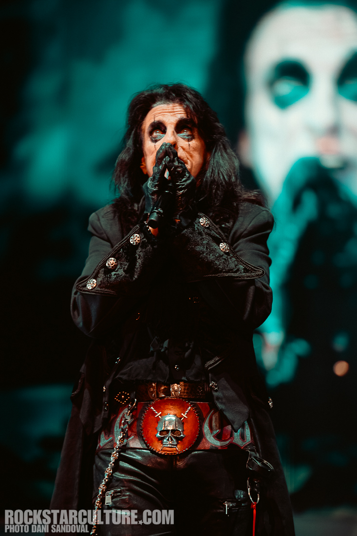 A performer wearing black attire with dramatic eye makeup, striking a pose on stage, with a skull belt and a digital backdrop.