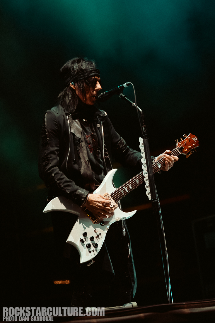 Musician performing on stage with a white electric guitar, wearing a black outfit and headband, microphone in front of them, with a moody green background.