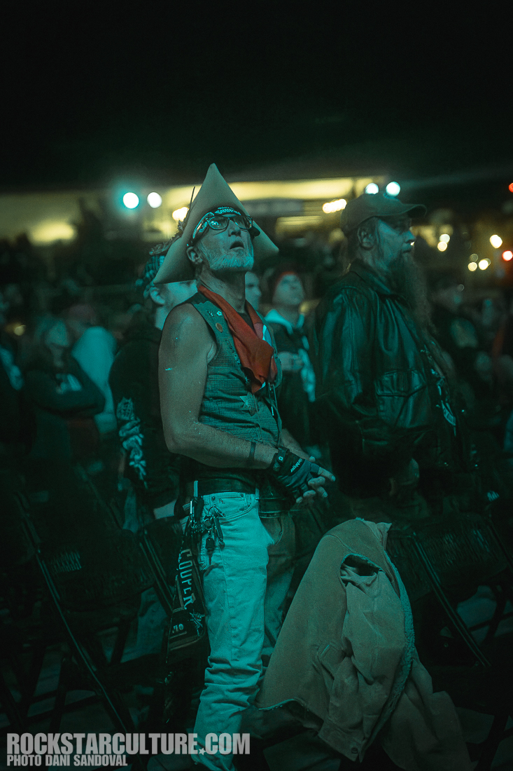 An enthusiastic concertgoer wearing a cowboy hat and a red bandana looks up, captivated by the performance, with a crowd visible in the background.