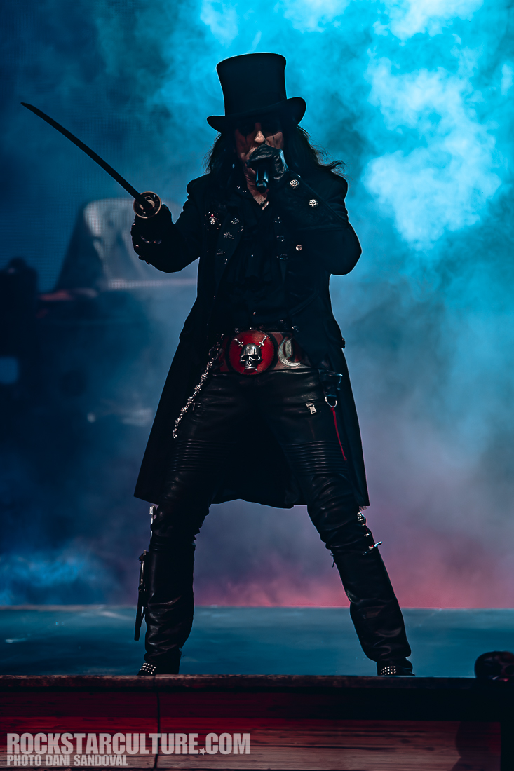 A performer in a black coat and top hat holds a microphone and a sword while standing on stage with smoke and colorful lighting in the background.