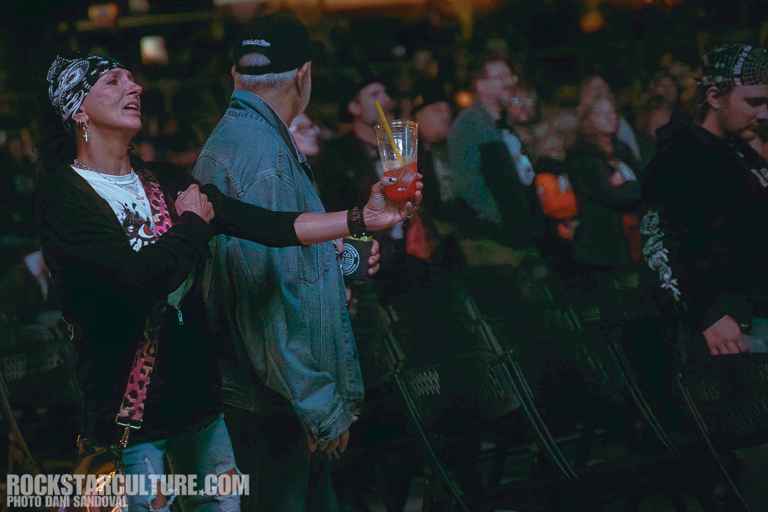 A woman wearing a bandana and black clothing holds a drink while interacting with a man in a denim jacket at a concert, with a crowd in the background.