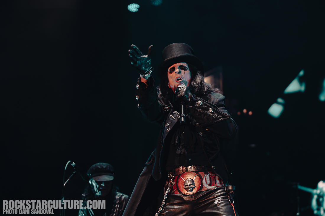 A performer in a black and red outfit, wearing a top hat, passionately singing into a microphone on stage. The background features dim lighting and other musicians. The scene conveys a vibrant concert atmosphere.