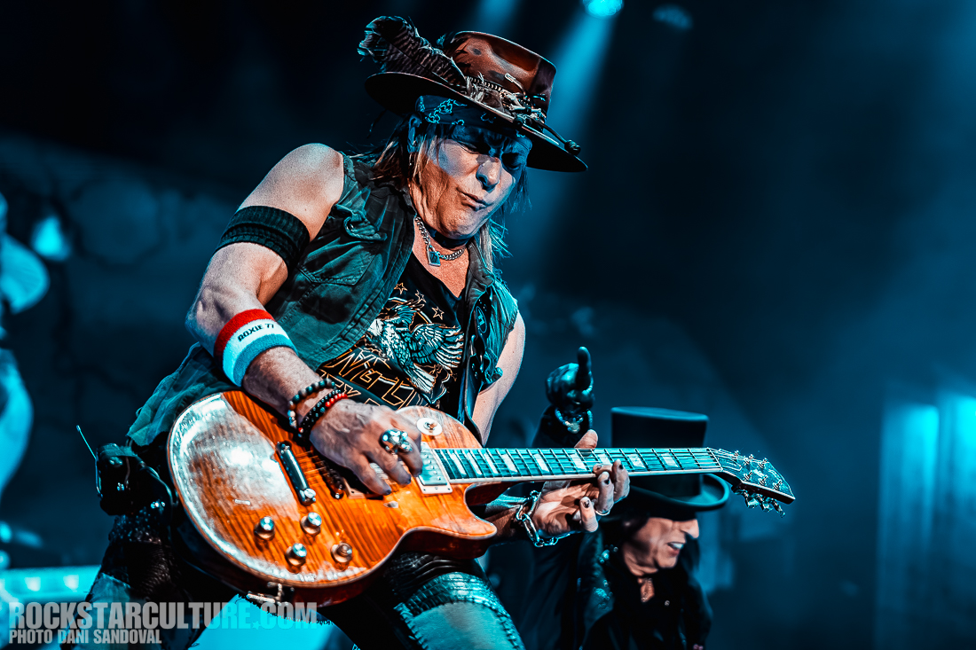 A guitarist performing on stage, wearing a black vest and hat, with expressive facial features, playing an electric guitar under colorful stage lights.