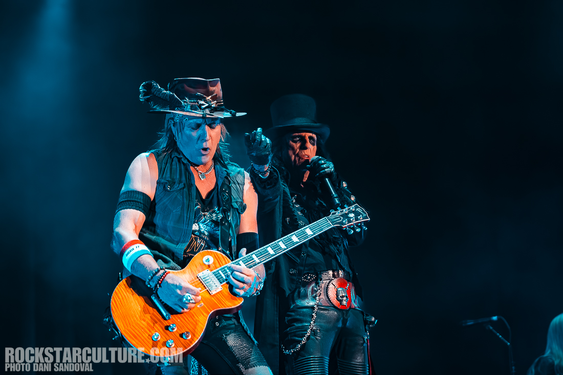 Two male musicians performing on stage; one playing an electric guitar and the other singing into a microphone, both wearing distinctive costumes and accessories.