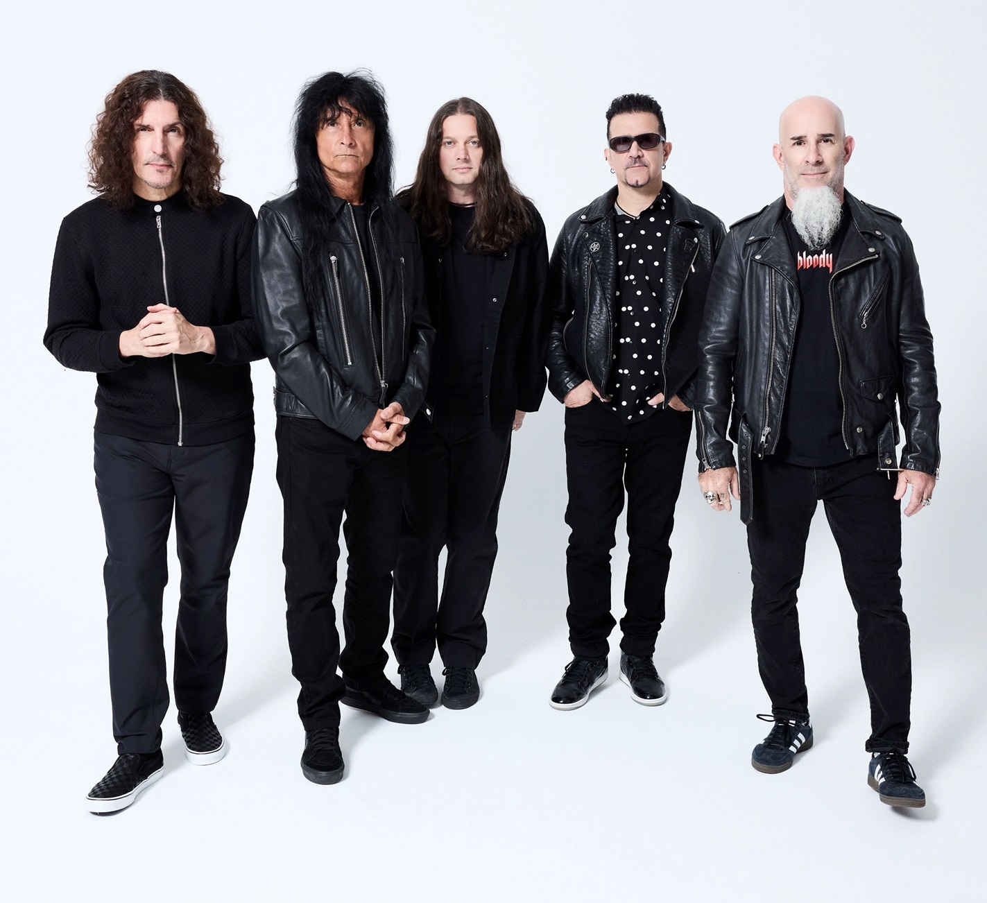 Five musicians standing together, dressed in black clothing and leather jackets, with varying hairstyles and an expressive group pose.