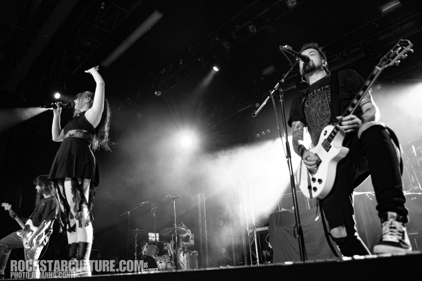 A black and white image of a live band performance with a female singer raising her right arm, and a male guitarist singing into a microphone. The background features other band members and stage lights.