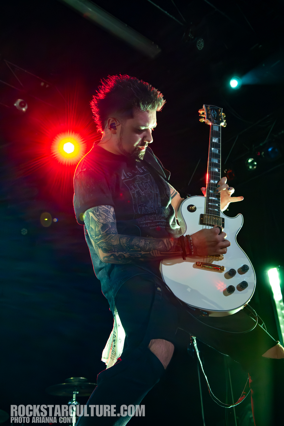 A male guitarist with a mohawk hairstyle performs on stage, holding a white electric guitar, illuminated by vibrant stage lights.