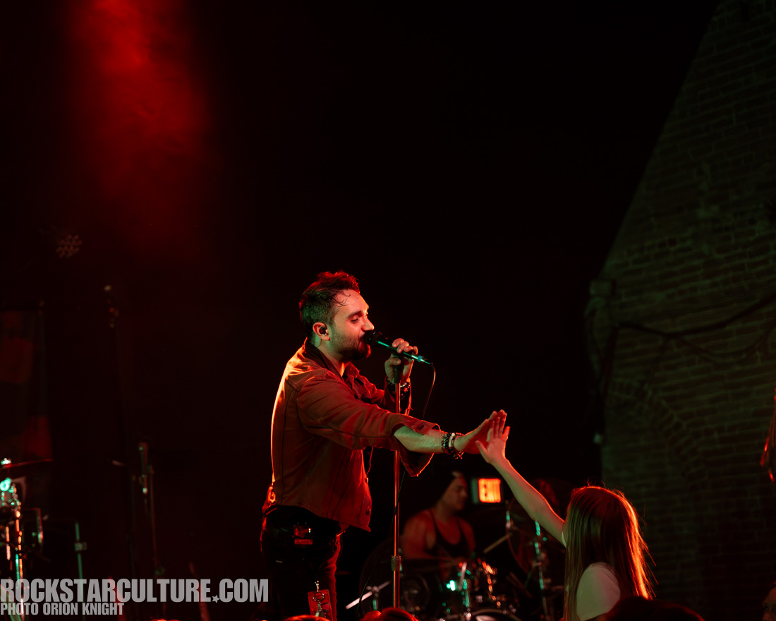 A male singer performing on stage, reaching out to a fan in the audience while holding a microphone, with a band member visible in the background.