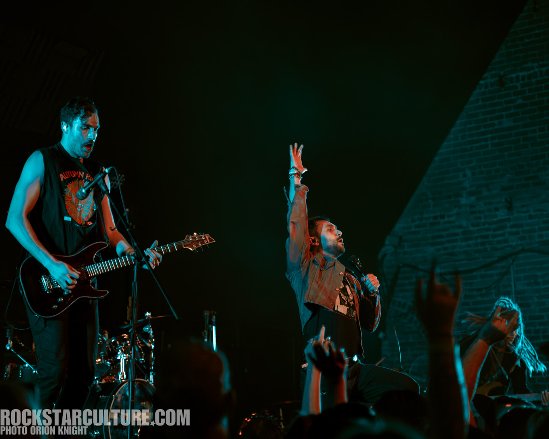 A live music performance featuring a vocalist passionately singing into a microphone with his hand raised, accompanied by a guitarist playing an electric guitar on stage.