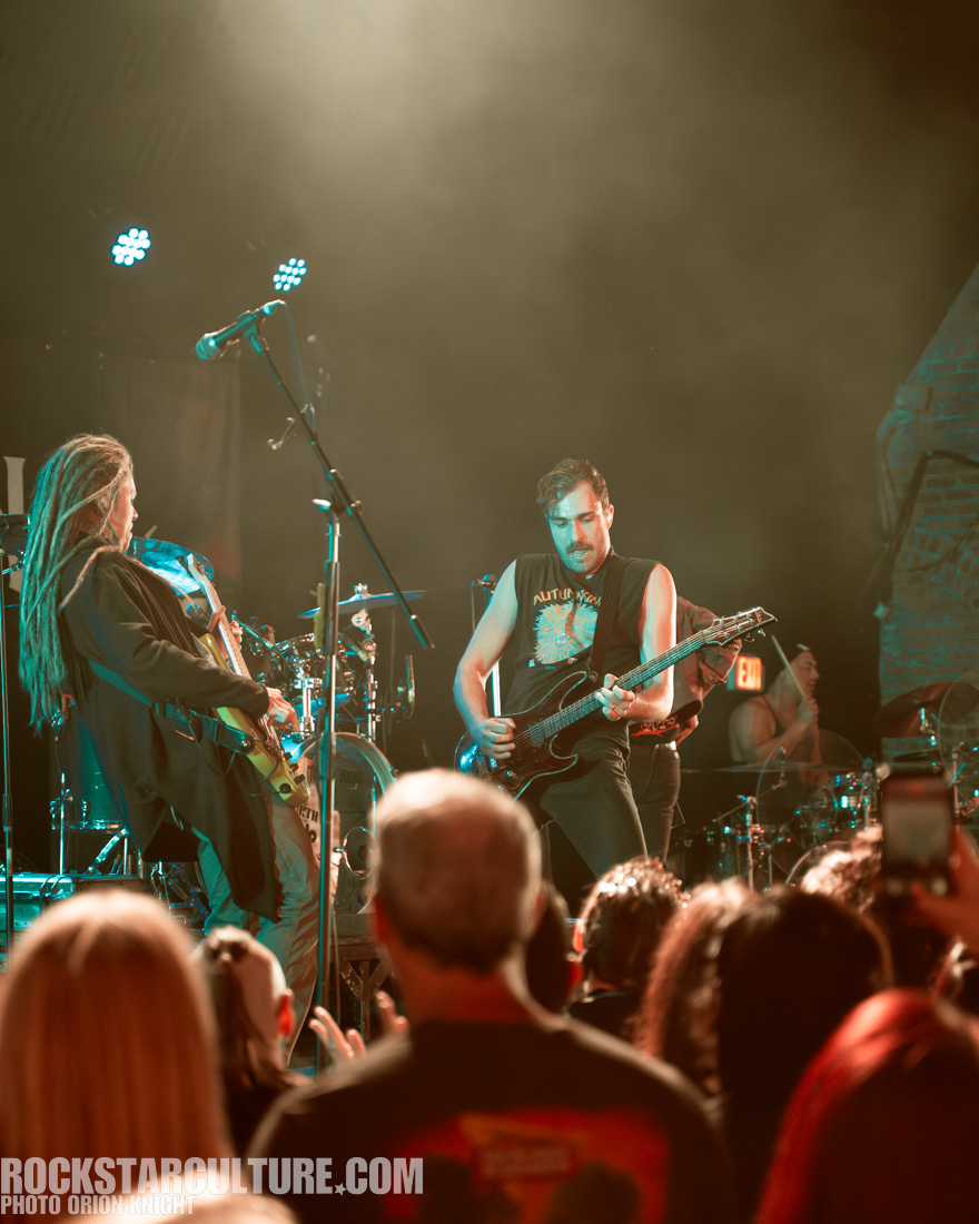 A live music performance featuring two guitarists on stage, with one musician playing an electric guitar and the other with dreadlocks playing a guitar. The audience is visible in the foreground, enjoying the performance.
