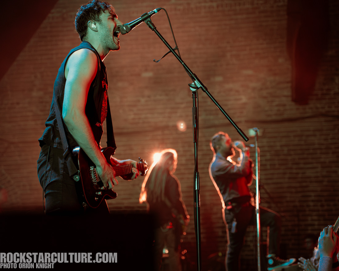 A male guitarist passionately performing on stage, singing into a microphone, with vibrant lighting in the background. Another vocalist is seen behind him, engaging with the audience.