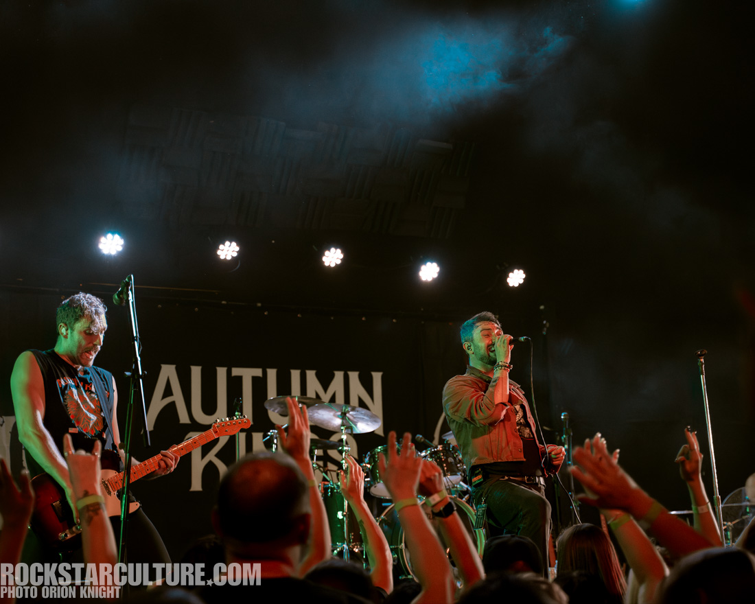 A live band performance featuring a singer holding a microphone and a guitarist playing an electric guitar on stage. The audience in the foreground is cheering with hands raised. The backdrop displays the band's name, 'Autumn King', under dramatic lighting.