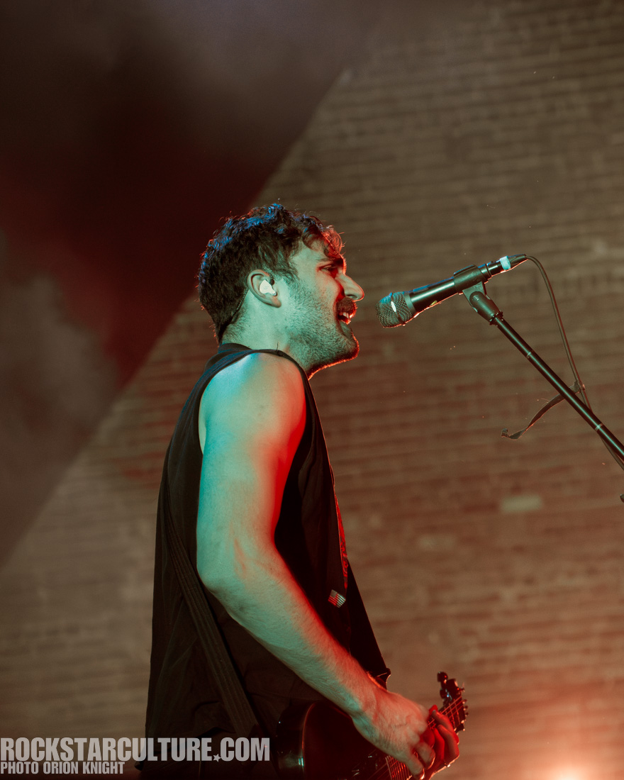 A musician performing on stage, singing into a microphone, with colorful lighting in the background.
