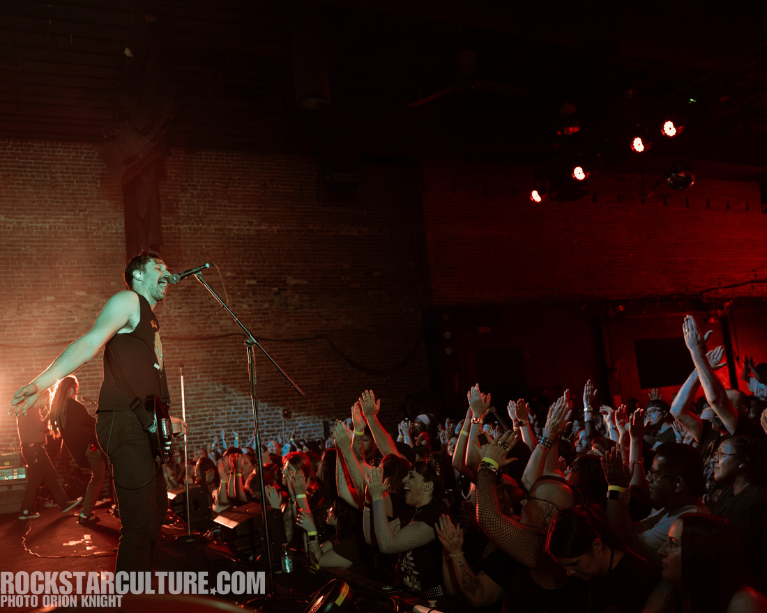 A live concert scene featuring a male vocalist singing passionately into a microphone, with a guitarist in the background. The audience is energetically applauding, with hands raised in excitement. The venue has a brick wall and colorful stage lights creating a vibrant atmosphere.