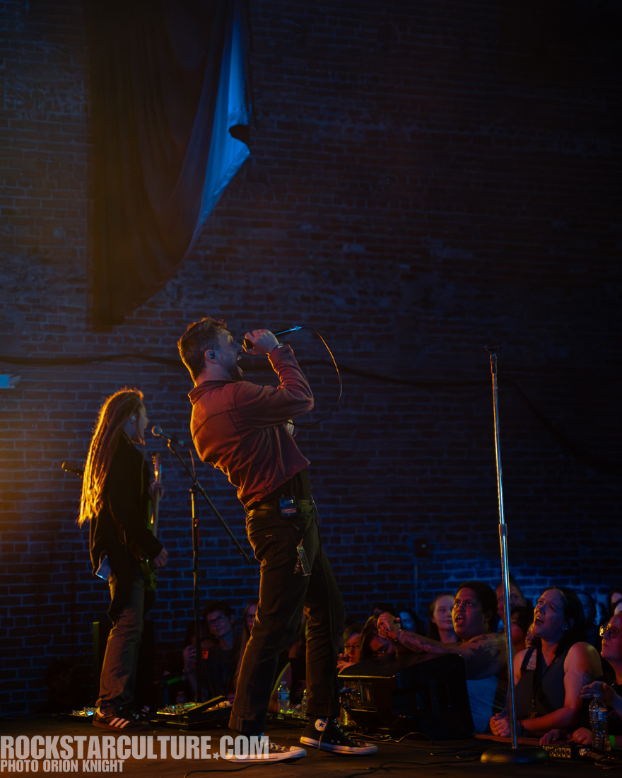 A male singer performing passionately on stage with a microphone, accompanied by another musician in the background. The audience is engaged, with some fans visibly enjoying the performance.