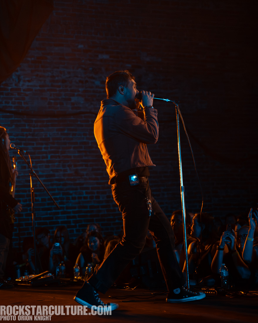 A male singer performing on stage, passionately singing into a microphone with a spotlight on him. He is wearing a jacket and standing in front of an audience who are holding up their phones to capture the moment. The backdrop features a textured brick wall, illuminated with blue and orange lighting.