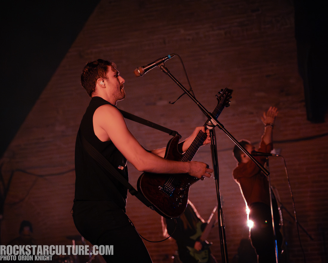 A musician playing guitar on stage during a performance, with dynamic lighting and a blurred band member in the background.