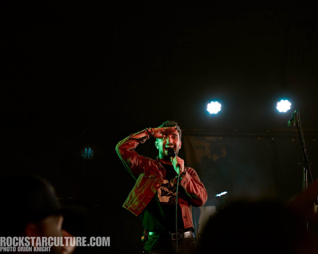 A male performer on stage salutes while holding a microphone, wearing a red jacket and black shirt, with bright stage lights in the background.