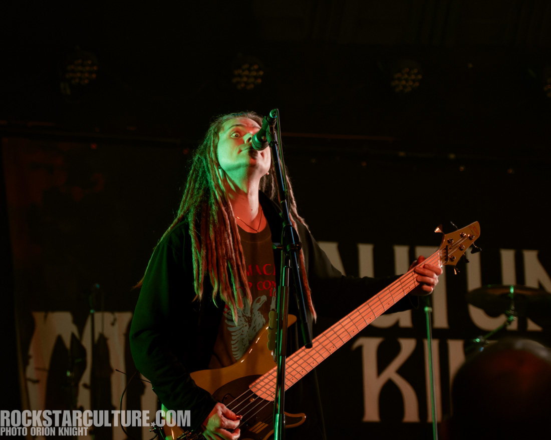 Musician performing on stage with a bass guitar and microphone, wearing a black hoodie and a graphic t-shirt, with dreadlocks and dramatic lighting.