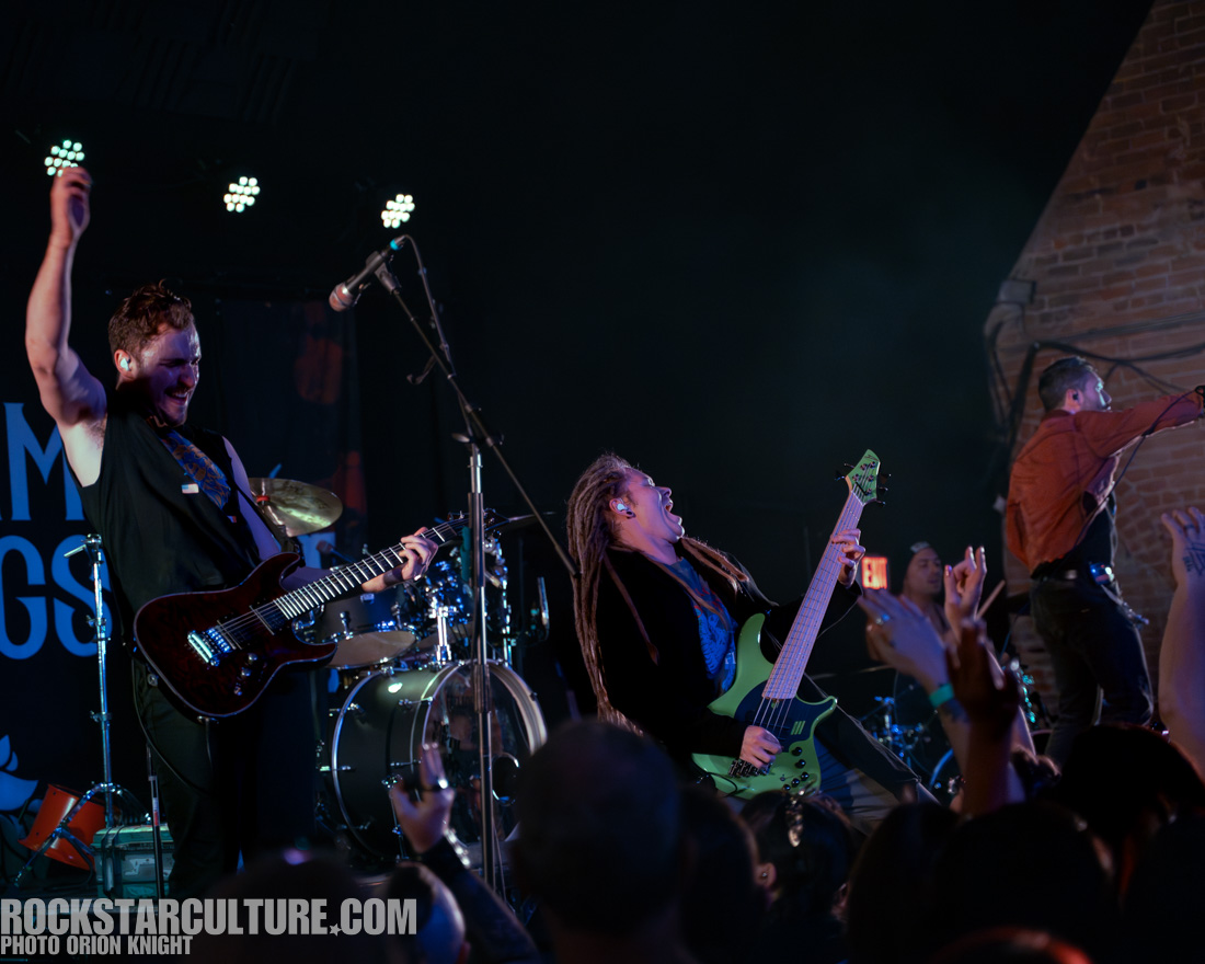 A lively concert scene featuring a band performing on stage. The guitarist on the left is enthusiastically playing while raising his arm, and the bassist on the right is passionately rocking out with a green bass guitar. The backdrop includes drums and a vibrant crowd enjoying the performance.