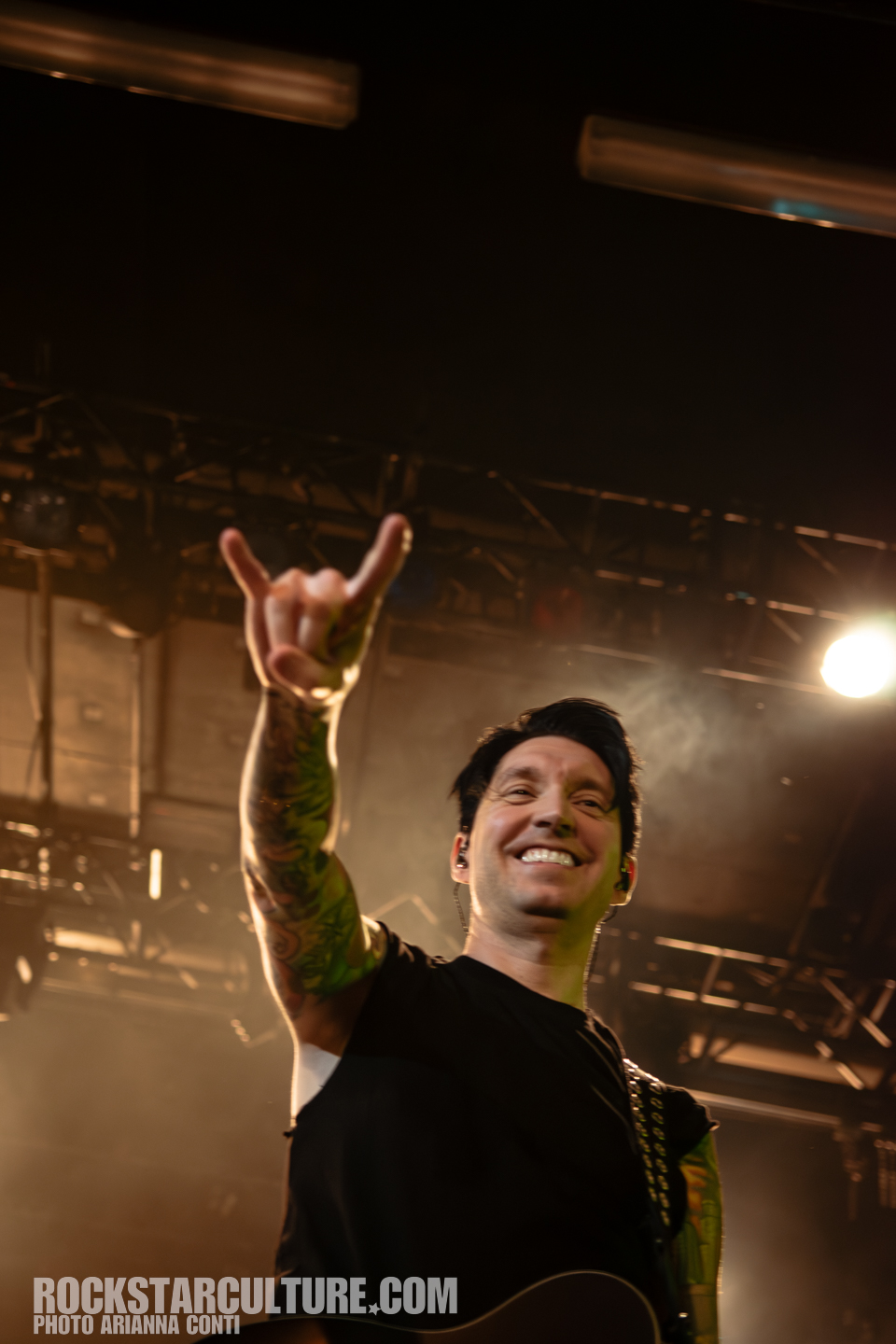 A smiling musician on stage gives a 'rock on' hand gesture while holding a guitar, with dramatic lighting and a blurred audience in the background.