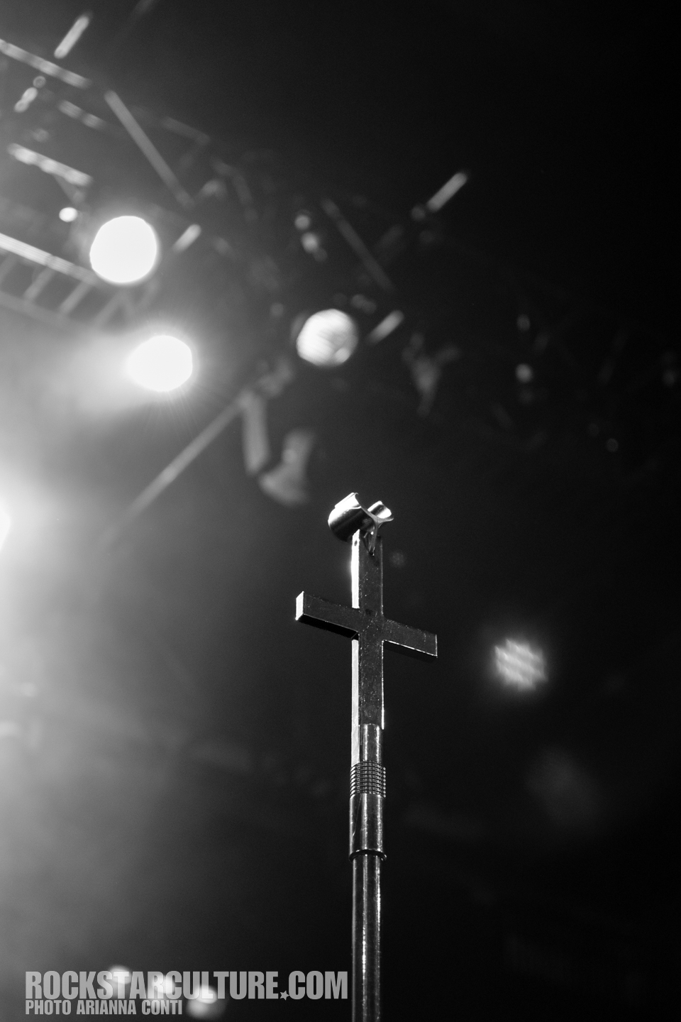 Close-up of a microphone stand with a cross at the top, against a blurred background of stage lights in black and white.