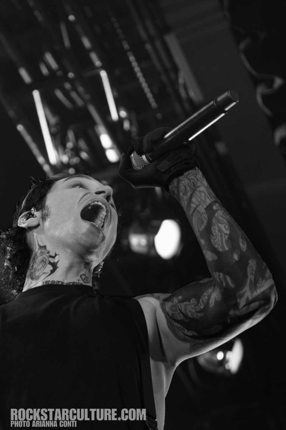 Black and white image of a male vocalist passionately singing into a microphone, showcasing detailed tattoos on his arms.