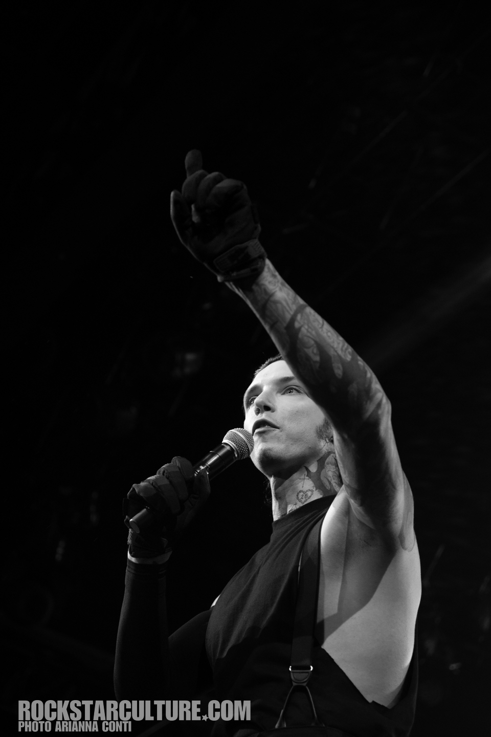 Black and white image of a male singer on stage, holding a microphone and gesturing passionately with his right hand, wearing gloves and a sleeveless shirt.