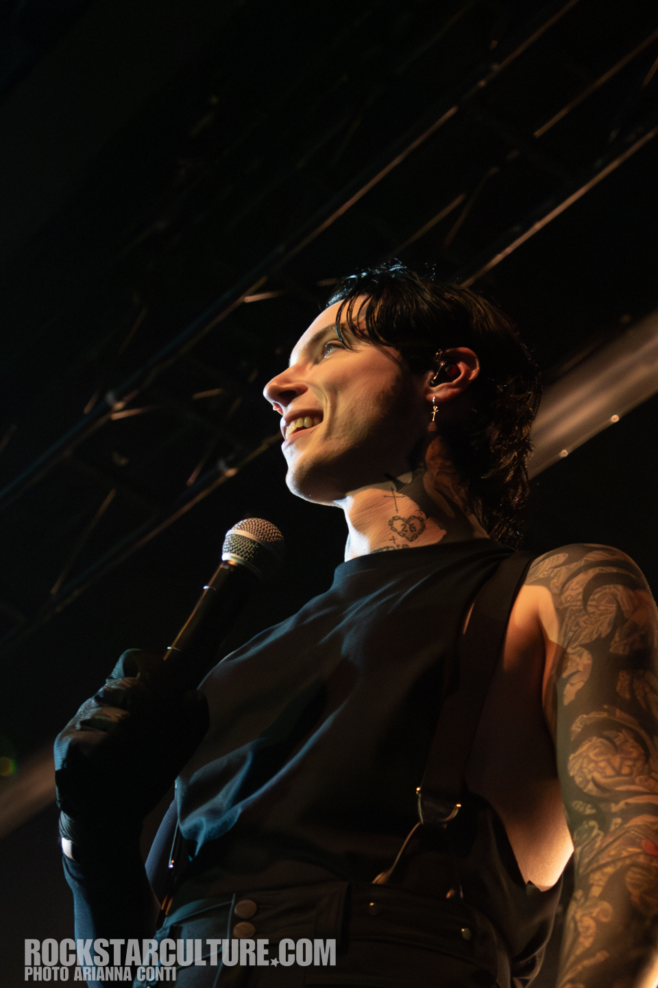 A performer with tattoos and earrings smiles while holding a microphone on stage. The background shows a stage setting with lights.