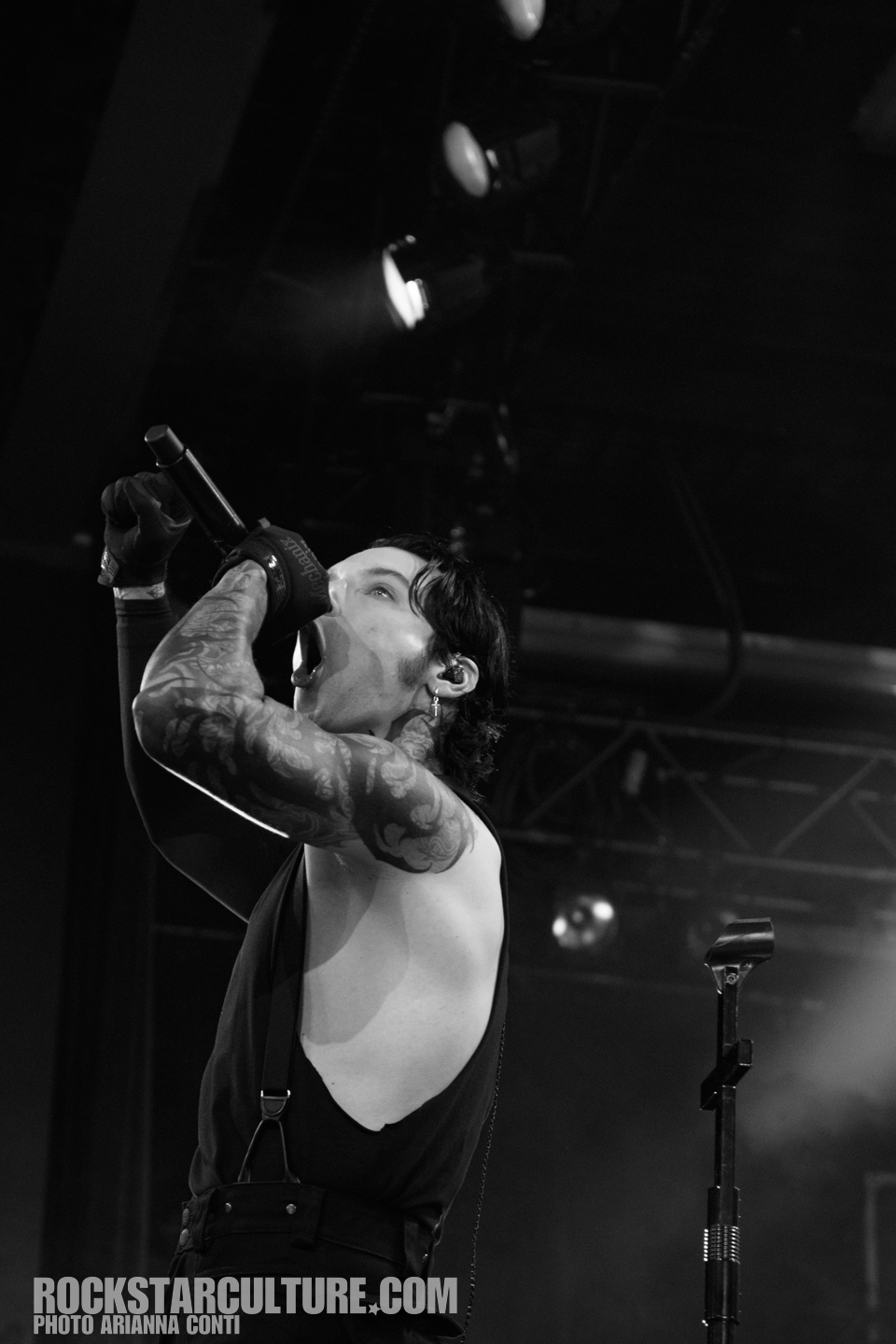 A male musician with tattoos passionately singing into a microphone on stage, wearing a sleeveless shirt and gloves, captured in black and white.