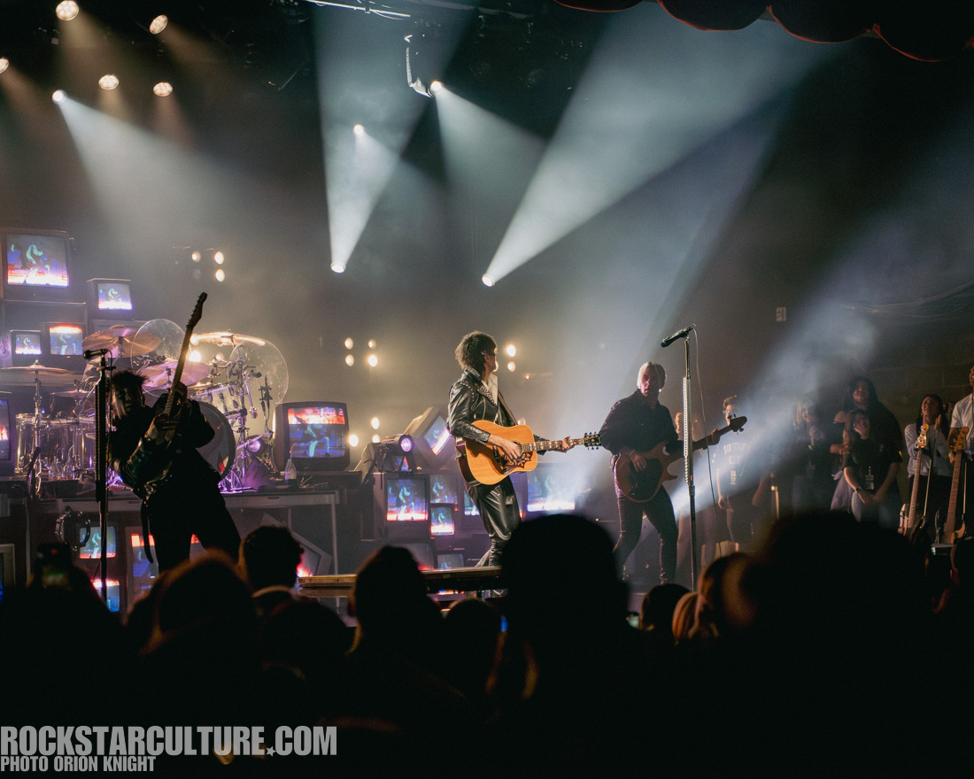 A live band performing on stage with dramatic lighting, featuring guitarists and a vocalist holding an acoustic guitar, surrounded by visible musical instruments and monitors displaying visuals.