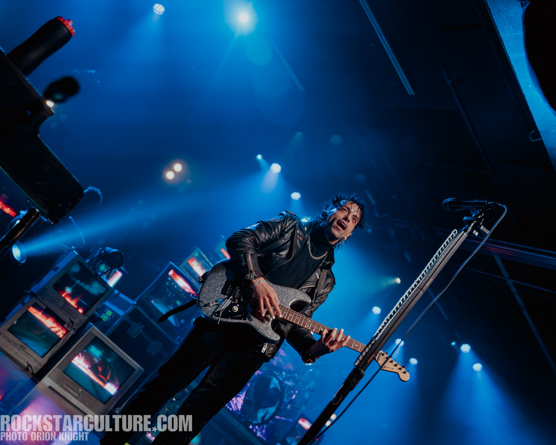 A musician performing on stage, holding a sparkling electric guitar, with vibrant blue lighting and retro televisions in the background.