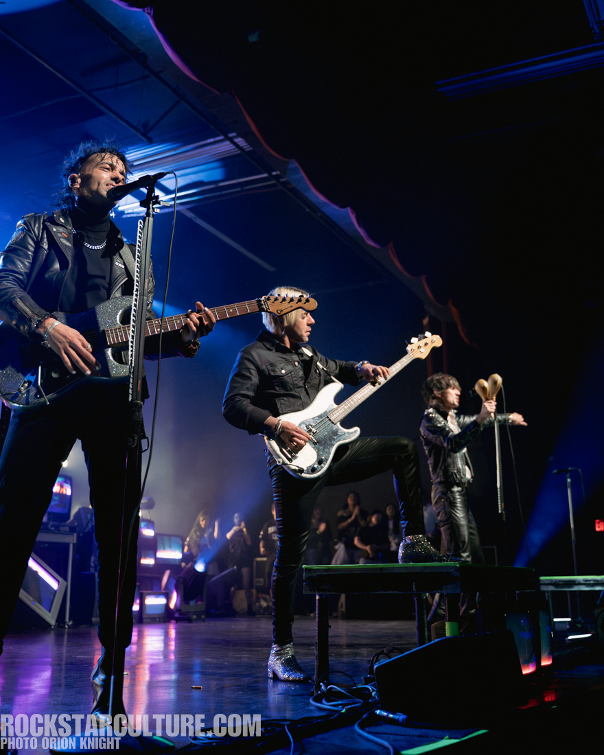 Three musicians performing on stage with electric guitars, surrounded by colorful stage lighting.