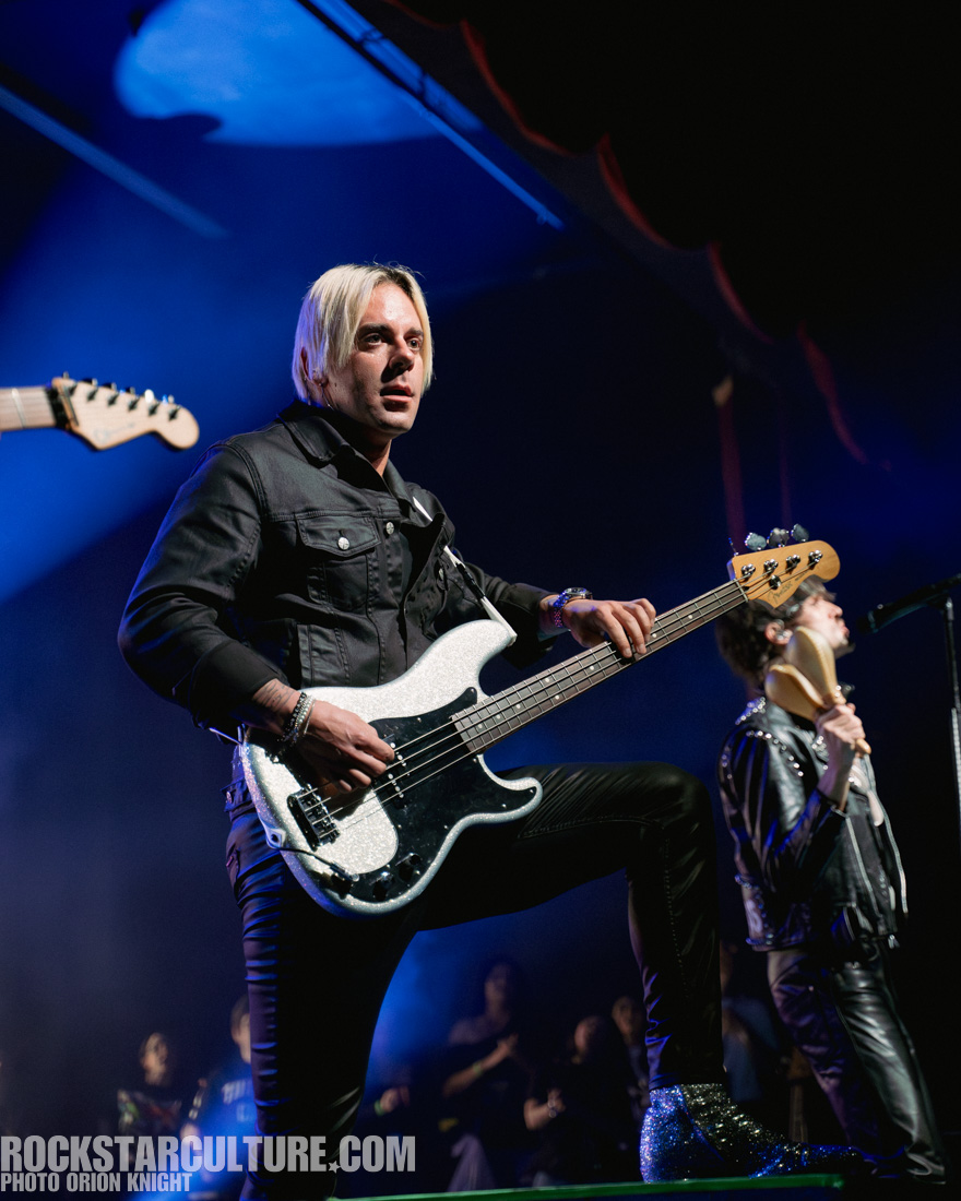A male bassist performing on stage with a glittery white bass guitar. He has blonde hair and is wearing a black jacket and pants. The background features blue stage lighting and other band members.